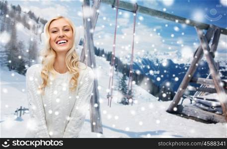 winter, vacation, christmas and people concept - smiling young woman in earmuffs and sweater over snowy mountains and wooden swing background