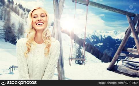 winter, vacation, christmas and people concept - smiling young woman in earmuffs and sweater over snowy mountains and wooden swing background. smiling young woman in winter earmuffs and sweater