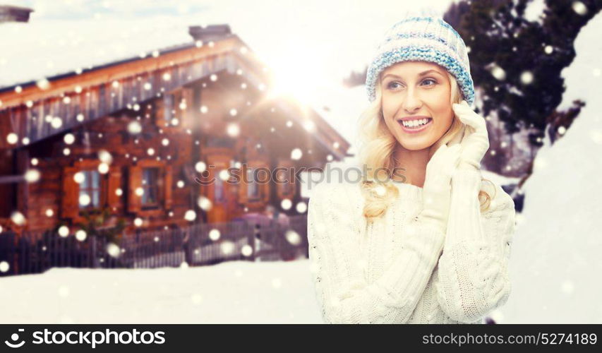 winter, vacation, christmas and people concept - smiling young woman in hat, sweater and gloves over wooden country house and snowflakes background. smiling young woman in winter hat and sweater