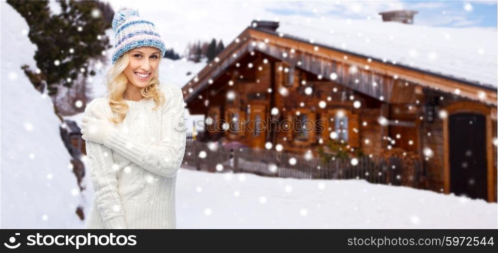 winter, vacation, christmas and people concept - smiling young woman in hat, sweater and gloves over wooden country house and snowflakes background