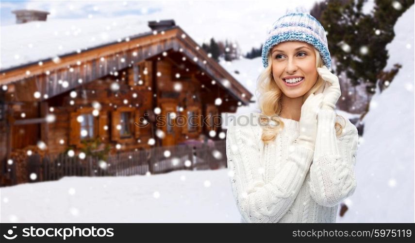winter, vacation, christmas and people concept - smiling young woman in hat, sweater and gloves over wooden country house and snowflakes background