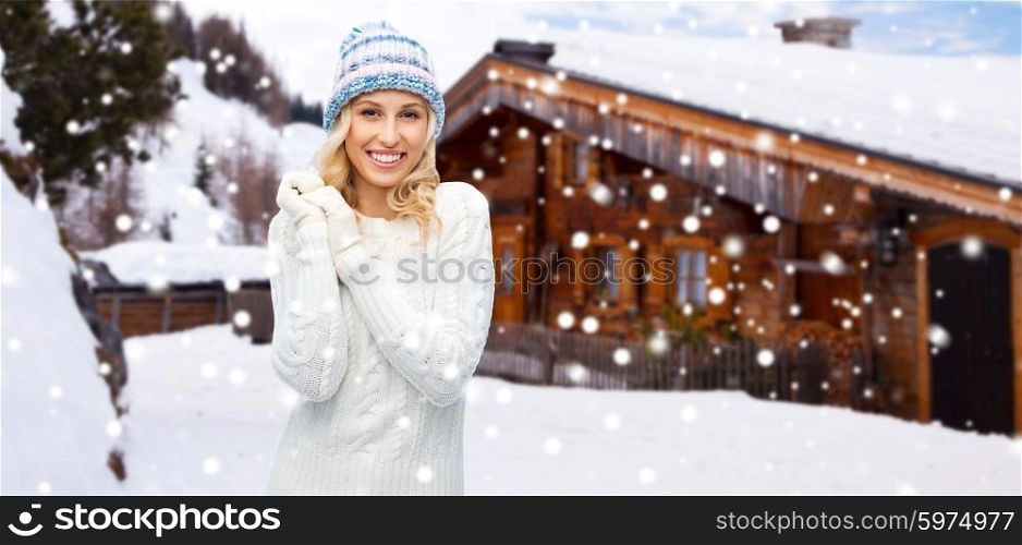 winter, vacation, christmas and people concept - smiling young woman in winter hat, sweater and gloves over wooden country house and snow background