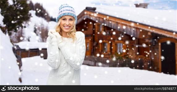 winter, vacation, christmas and people concept - smiling young woman in winter hat, sweater and gloves over wooden country house and snow background