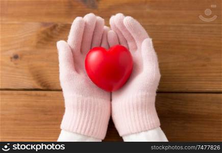 winter, valentine&rsquo;s day and christmas concept - hands in pale pink woollen gloves holding red heart over wooden boards background. hands in pink woollen gloves holding red heart