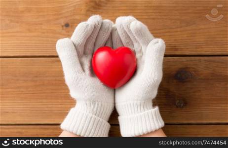 winter, valentine&rsquo;s day and christmas concept - hands in white woollen gloves holding red heart over wooden boards background. hands in white woollen gloves holding red heart