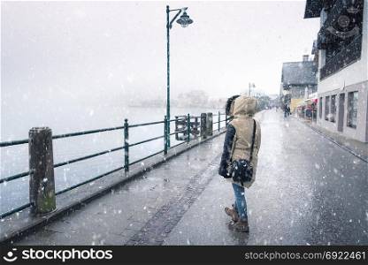 Winter weather theme image with a woman dressed for cold, walking on the street, under the snowfall, near the Hallstatter lake, in Hallstatt town, Austria.
