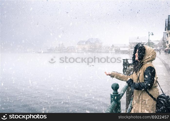 Winter weather theme image with a young woman smiling and holding hand to catch snowflakes, by the Hallstatter lakeshore, in Hallstatt village, Austria.