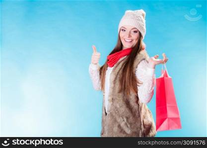 Winter woman in warm clothing with red paper shopping bag showing thumb up on blue. Girl buying. Sale, consumerism and retail.