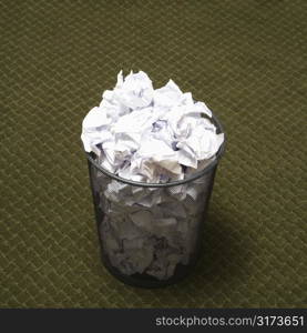 Wire mesh trash can filled with crumpled paper on green carpet.