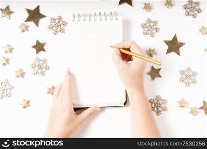 Wish list for Cristmas and New Year. Holiday decorations and ruled notebook with wish list and somwones hands, flat lay top view. Christmas flat lay scene with golden decorations