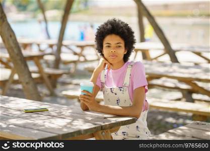 Wistful African American female with takeaway coffee looking away while sitting at wooden table in park on sunny summer day. Black woman with coffee in park