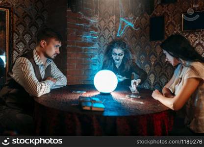 Witch reads a magic spell over a crystal ball, young couple on spiritual seance. Female foreteller calls the spirits