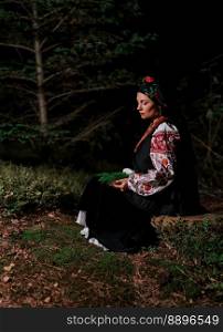 Witch woman collects herbs, ferns at night in Carpathian mountains forest. She in in traditional ukrainian handkerchief, national dress - vyshyvanka, ancient coral beads. Folk medicine concept.. Witch woman collects herbs, ferns at night in Carpathian mountains forest.