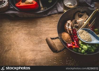 Wok pot with vegetarian asian cuisine ingredients for stir fry with chopped vegetables, spices, sesame seeds and lemongrass on rustic wooden background, top view. Chinese or Thai food cooking concept