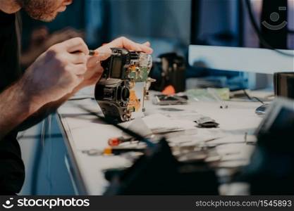 Woking process. Unknown man repairs professional digital camera, uses screwdriver, poses at workplace, holds photocamera disassembled part