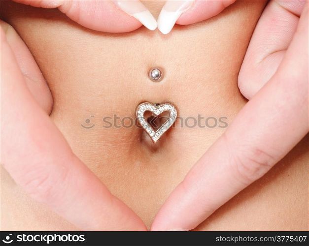 woman&#39;s hands forming a heart symbol around navel with piercing. Mother awaiting for the baby.