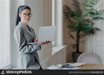 Woman administrative stands near desktop, holds modern laptop computer, uses wireless connection to internet, dressed in formal grey outfit, searches information on web page, works distantly