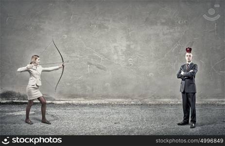 Woman aiming her goal. Young woman archer in suit aiming to shoot apple on man head