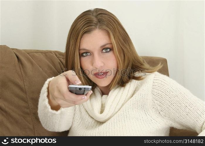 Woman aiming remote control towards camera. Sitting on couch. Wearing cream sweater.