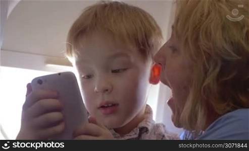 Woman and her grandchild in the plane. Child listening to grandmother and looking at smart phone screen