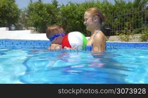 Woman and little boy are playing with inflatable ball in open-air swimming pool.