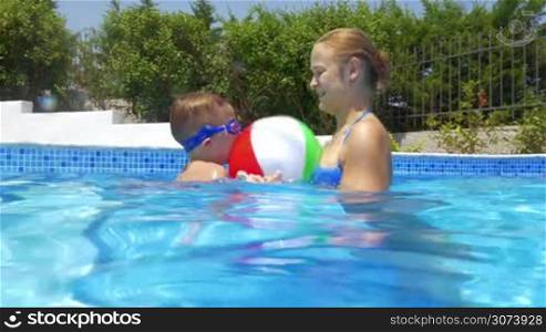 Woman and little boy are playing with inflatable ball in open-air swimming pool.