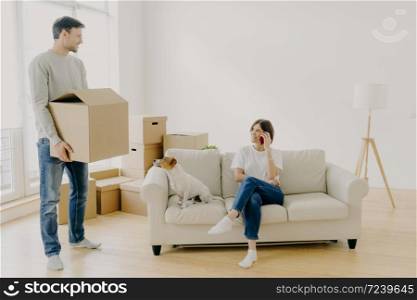 Woman and man real estate renters pose in empty living room, female sits on couch in middle of room, has phone conversation, share impressions after moving in new apartment, male carries boxes