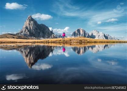 Woman and mountains reflected in lake at sunset in autumn in Dolomites, Italy. Standing girl on the shore of lake is looking on high rocks and blue sky with clouds in fall. Perfect reflection in water. Woman and mountains reflected in lake at sunset in autumn