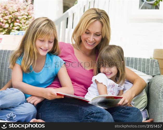 Woman and two young girls sitting on patio reading book smiling