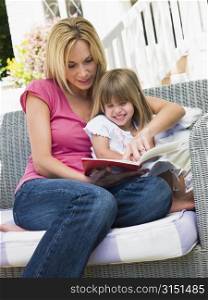 Woman and young girl sitting on patio reading book smiling