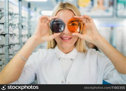 Woman applies lenses of different colors to the eyes, showcase with spectacles in optics store. Selection of eyeglasses with professional optometrist, sunglasses choice, eyecare concept