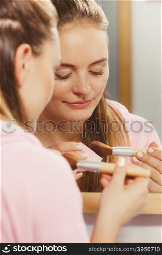 Woman applying bronzing powder with brush to her skin. Woman face painting. Girl applying rouge or bronzing powder with brush to her skin looking in bathroom mirror. Makeup cosmetics and beauty procedures.
