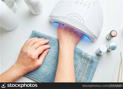Woman applying gel hybrid polish using UV lamp. Beauty wellness spa treatment concept. Cosmetic products, UV lamp, green leaves on white table. Spa, manicure, skin care concept. Flat lay, overhead view