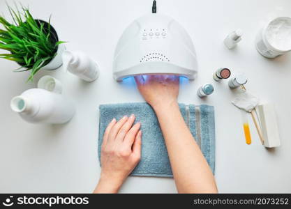 Woman applying gel hybrid polish using UV lamp. Beauty wellness spa treatment concept. Cosmetic products, UV lamp, green leaves on white table. Spa, manicure, skin care concept. Flat lay, overhead view