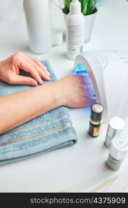 Woman applying gel hybrid polish using UV lamp. Beauty wellness spa treatment concept. Cosmetic products, UV lamp, green leaves on white table. Spa, manicure, skin care concept. Flat lay, overhead view