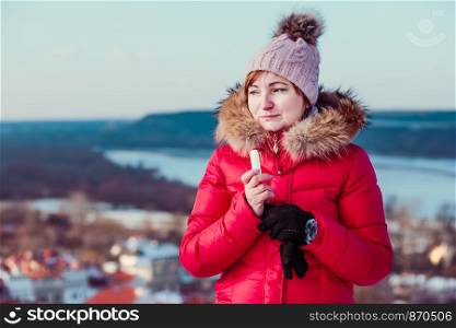 Woman applying lip balsam while walk on a wintery day. Wearing red coat and cap. Town in the background
