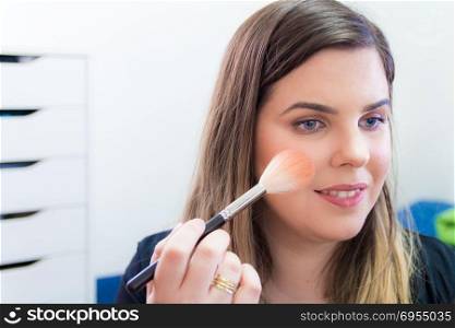 Woman applying makeup in her bedroom. She is smiling.