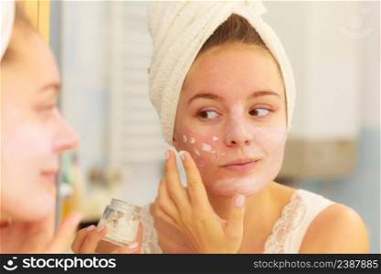 Woman applying mask moisturizing skin cream on face looking in bathroom mirror. Girl taking care of her complexion layering moisturizer. Skincare spa treatment. . Woman applying mask cream on face in bathroom