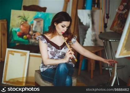 Woman artist painting a picture in a studio. Creative pensive painter girl paints a colorful picture on canvas with oil colors in workshop.
