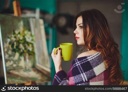 Woman artist painting a picture in a studio. Creative pensive painter girl paints a colorful picture on canvas with oil colors in workshop. Portrait of a beautiful woman wrapped in a plaid with a cup of tea