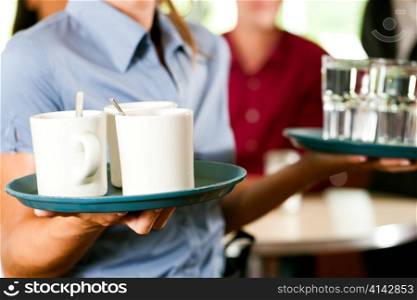 Woman as waitress in a bar or restaurant with coffee mugs; in the background are guests