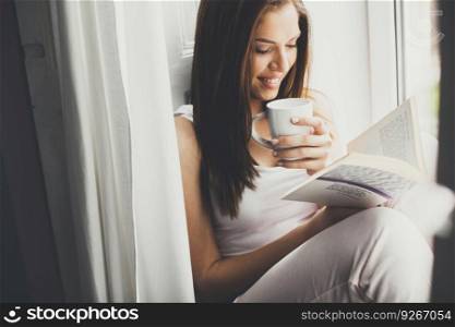 Woman at home sitting near window relaxing in her living room reading book and drinking coffee or tea