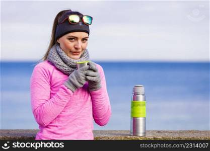 Woman athlete fit girl wearing warm sporty clothes outdoor in cold weather drinking hot tea from vacuum flask thermos, warming up. Sports, activities, hiking in winter or autumn time. Fit woman in cold day warming up drink tea