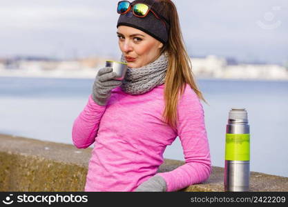 Woman athlete fit girl wearing warm sporty clothes outdoor in cold weather drinking hot tea from vacuum flask thermos, warming up. Sports, activities, hiking in winter or autumn time. Fit woman in cold day warming up drink tea