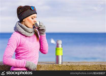 Woman athlete fit girl wearing warm sporty clothes outdoor in cold weather drinking hot tea from vacuum flask thermos, warming up. Sports, activities, hiking in winter or autumn time