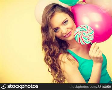Woman attractive cheerful girl holding colorful balloons and sweet lollipop in hands. Summer holidays, celebration and happiness concept. Studio shot bright yellow background. Woman with colorful balloons and lollipop