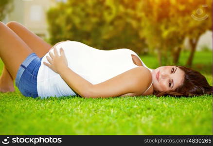 Woman awaiting for a baby, cute pregnant female lying down on fresh green grass in the garden, sunny day, happy and healthy pregnancy concept