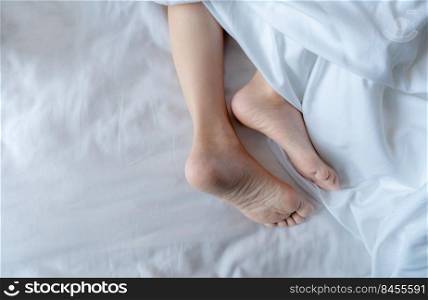 Woman barefoot on bed under white linen blanket in hotel or home bedroom. Healthy sleep and relaxation concept. Lazy Sunday morning. Bare feet of woman chilling sleep on white comfort bed and duvet.