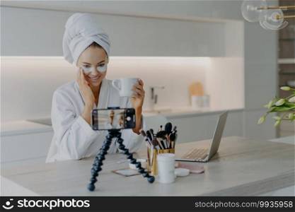 Woman beauty blogger shoots cosmetic vlog looks at camera of smartphone talks with followers applies collagen patches under eyes drinks tea wears white soft bathrobe records video broadcast.