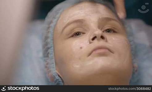 Woman being prepped in a skincare clinic as she lies on a couch in a surgical cap while the technician applies protective gel or cream to her face before the procedure